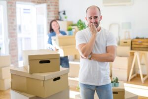 Young couple arround cardboard boxes moving to a new house, bald man standing at home looking stressed and nervous with hands on mouth biting nails.